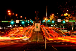 Night traffic intersection with red vehicle light trails, illuminated street signs, and photo enforcement sign at urban crossroads