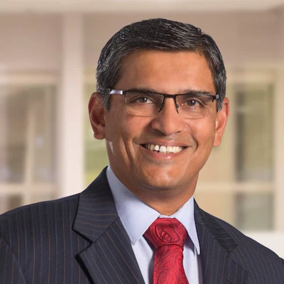 Professional headshot of man wearing dark pinstriped suit, light blue shirt, and red tie with glasses, smiling at camera
