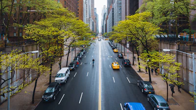 Tree-lined urban avenue with yellow taxis, parked cars, and pedestrians between tall buildings on a sunny day