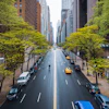 Tree-lined urban avenue with yellow taxis, parked cars, and pedestrians between tall buildings on a sunny day