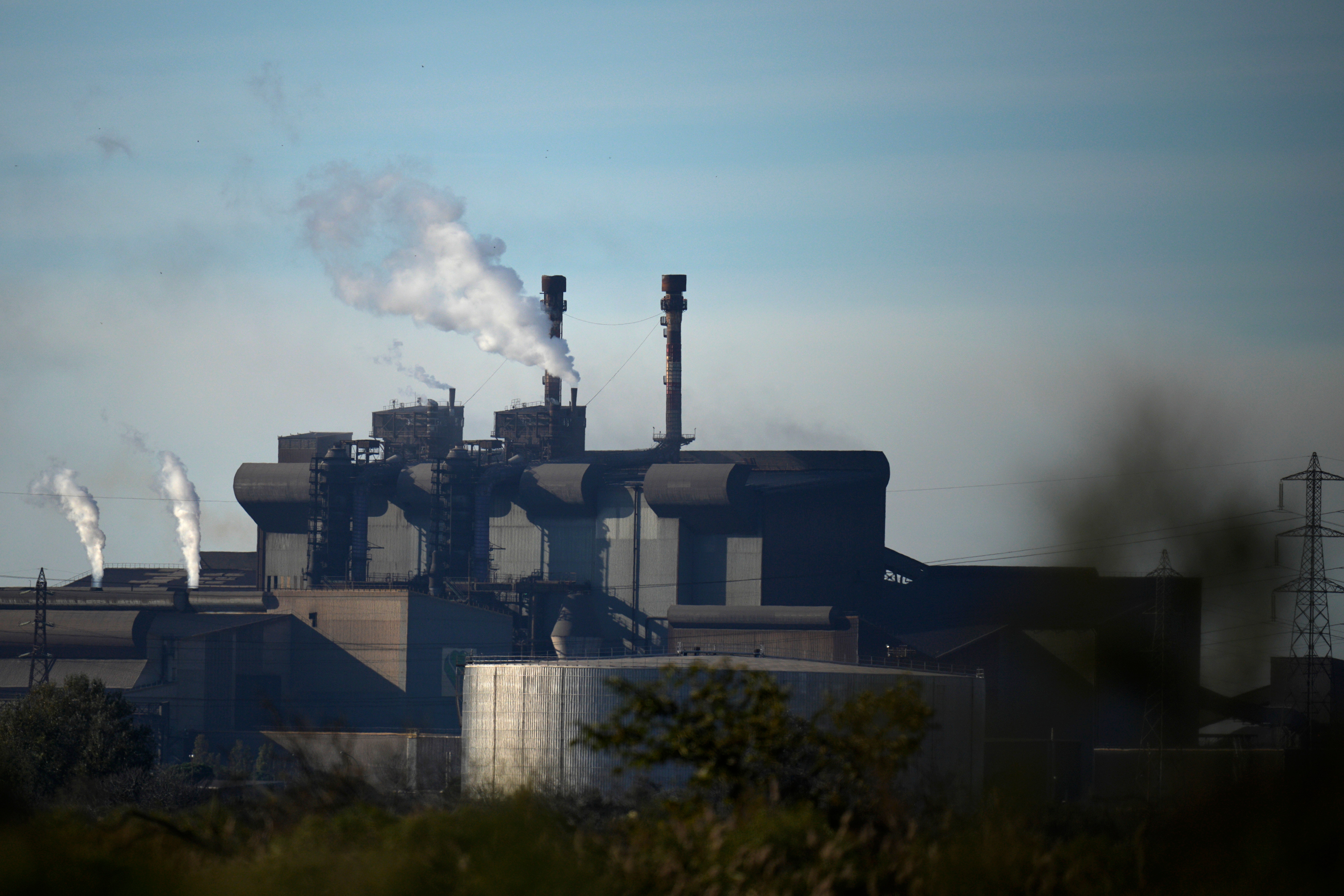 Industrial steel mill facility with multiple smokestacks emitting steam against a blue sky