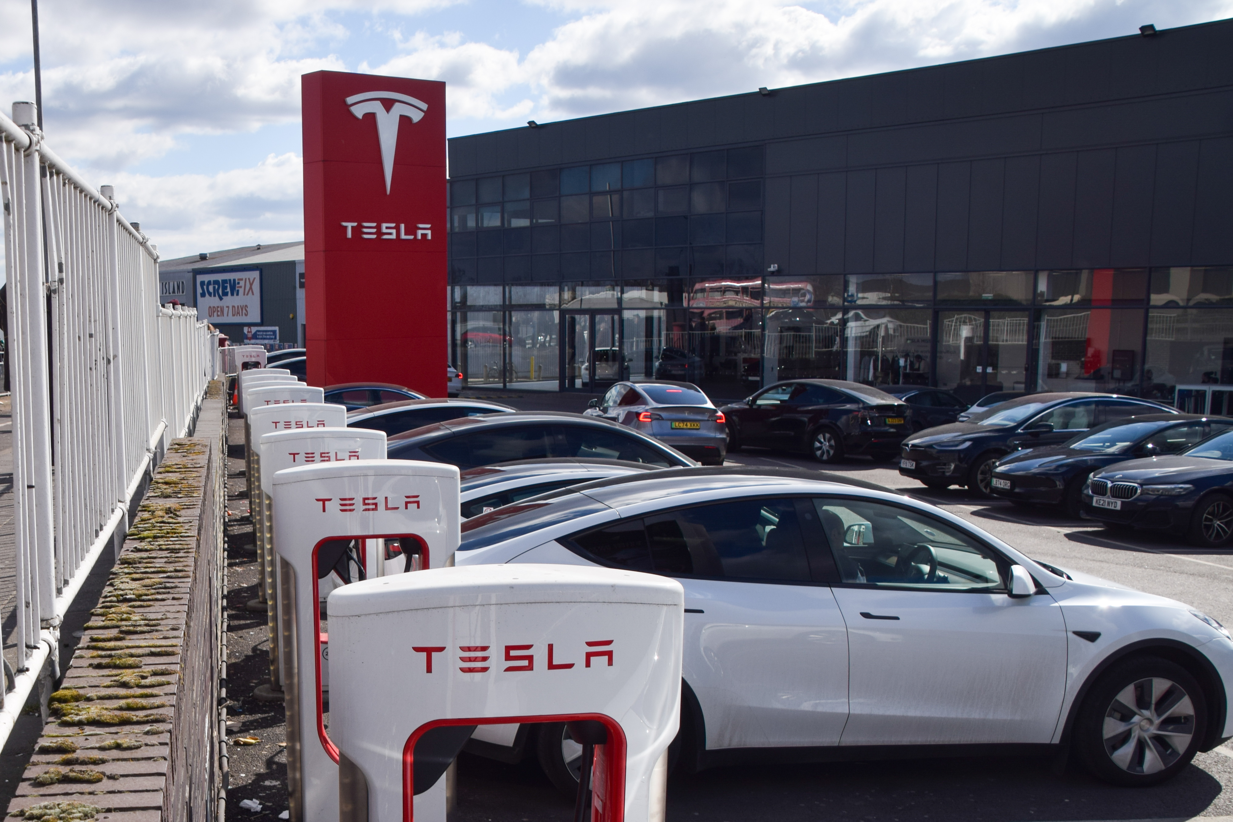 Tesla Supercharger station with white charging units and red branded signage at dealership facility with vehicles parked outside
