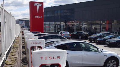 Tesla Supercharger station with white charging units and red branded signage at dealership facility with vehicles parked outside