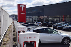Tesla Supercharger station with white charging units and red branded signage at dealership facility with vehicles parked outside