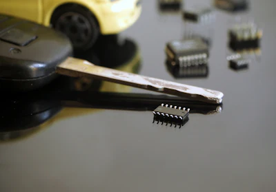 Tweezers holding integrated circuit chip with toy car and electronic components on gray surface