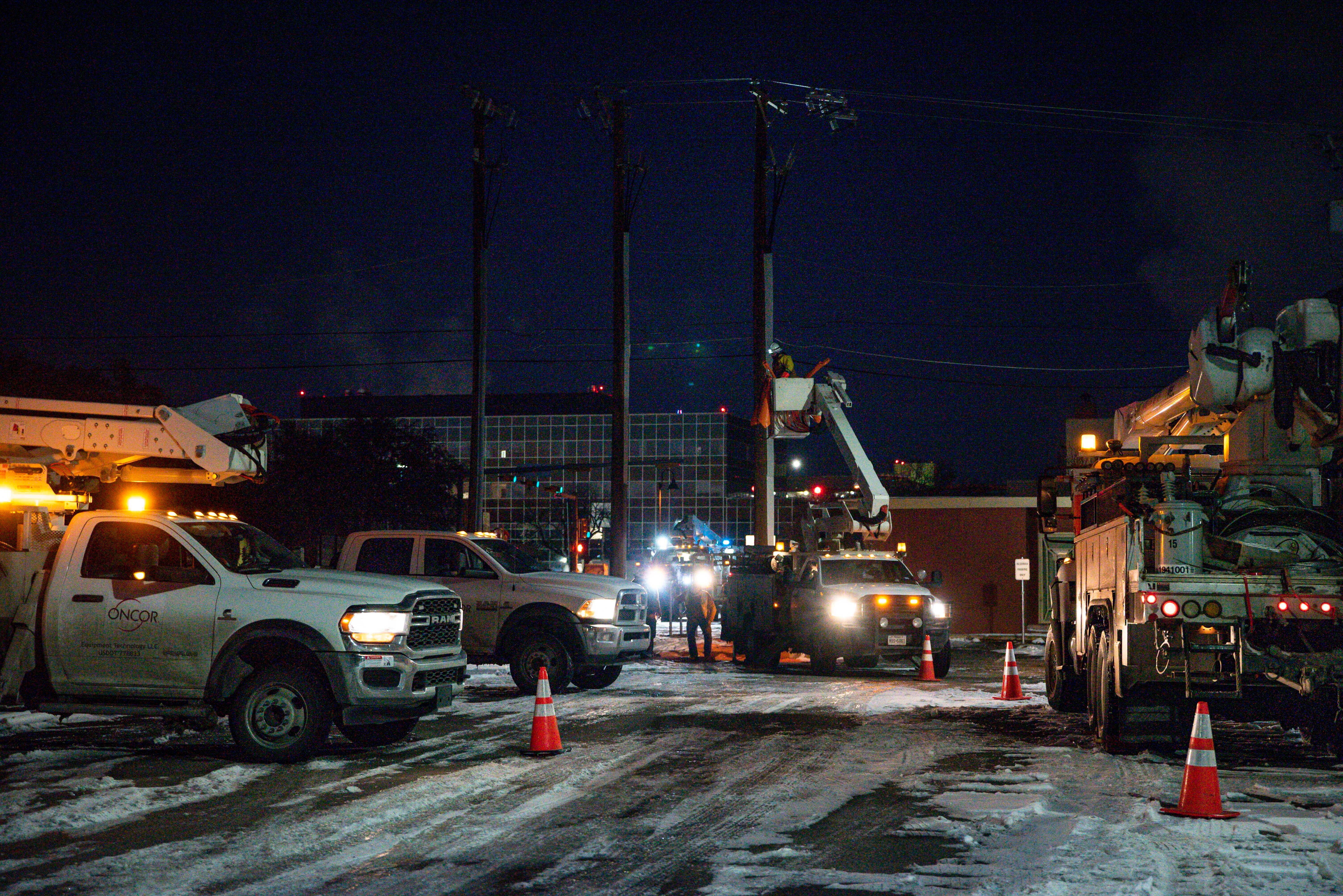 Nighttime utility work scene with service trucks, bucket lifts, and workers near power lines and industrial buildings.