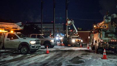 Nighttime utility work scene with service trucks, bucket lifts, and workers near power lines and industrial buildings.