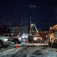 Nighttime utility work scene with service trucks, bucket lifts, and workers near power lines and industrial buildings.