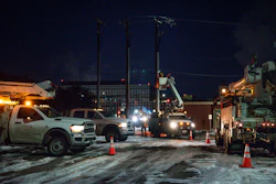 Nighttime utility work scene with service trucks, bucket lifts, and workers near power lines and industrial buildings.
