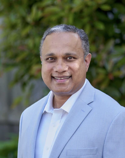 Professional headshot of a man in light blue blazer smiling outdoors with green foliage background