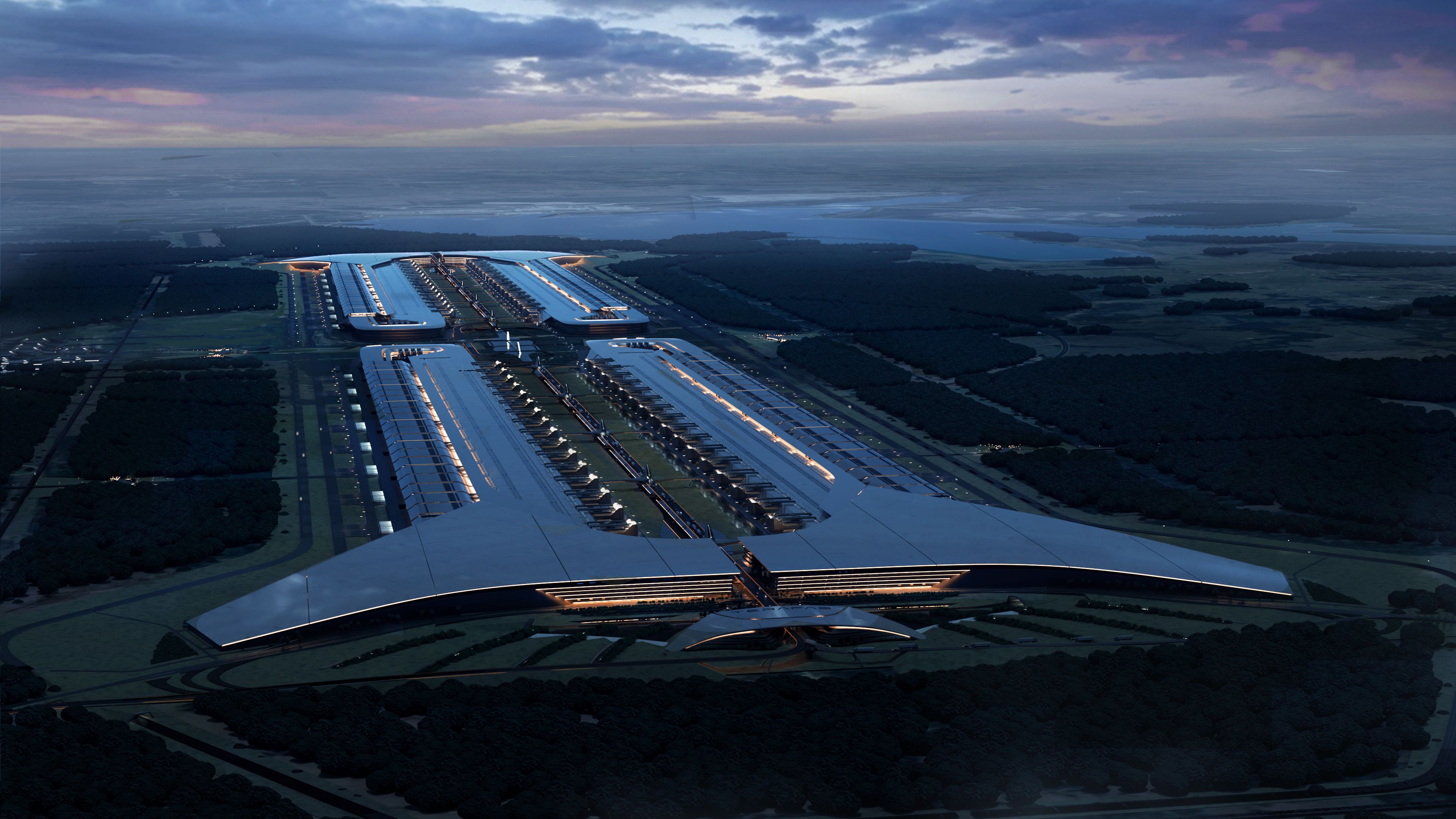 Aerial view of large industrial facility with parallel illuminated sections at twilight, surrounded by forests and fields