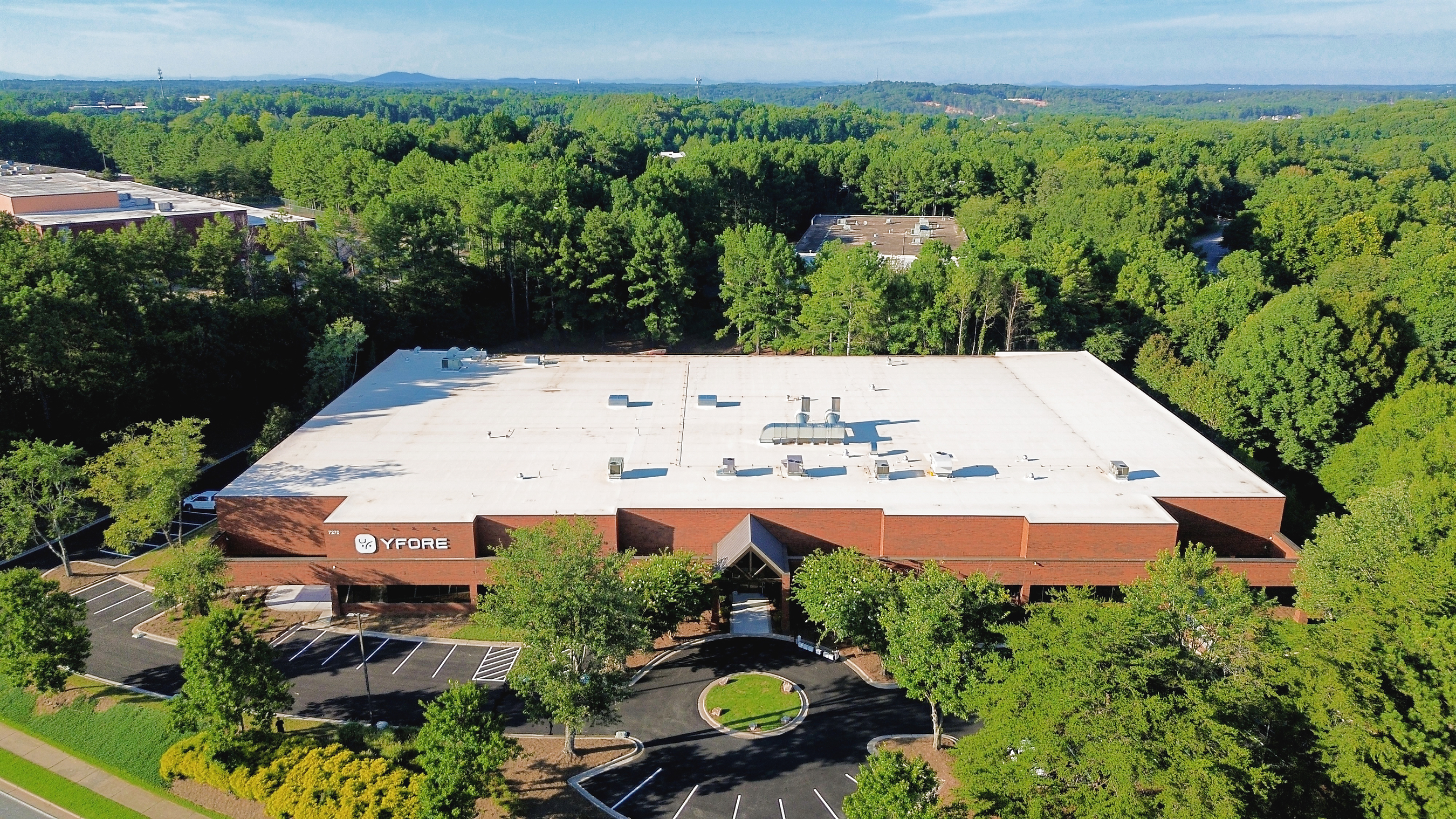Aerial view of modern industrial building with rust-colored exterior surrounded by dense green forest and parking areas