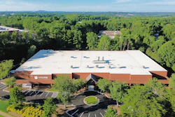 Aerial view of modern industrial building with rust-colored exterior surrounded by dense green forest and parking areas