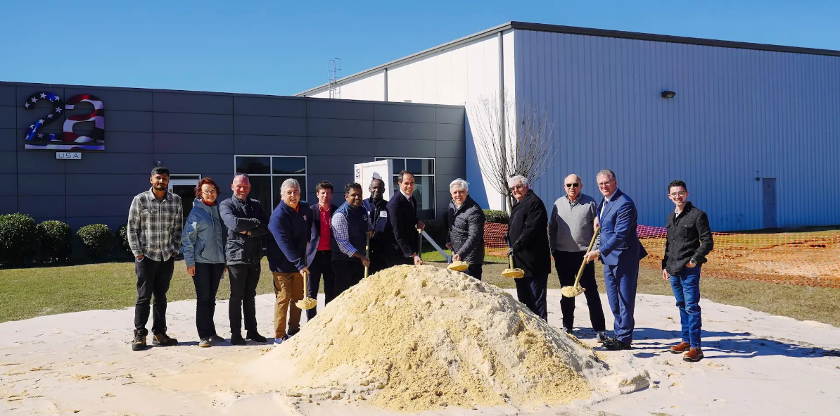 Group of people at groundbreaking ceremony standing around dirt pile in front of industrial building