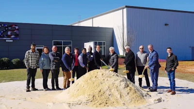Group of people at groundbreaking ceremony standing around dirt pile in front of industrial building