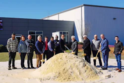 Group of people at groundbreaking ceremony standing around dirt pile in front of industrial building