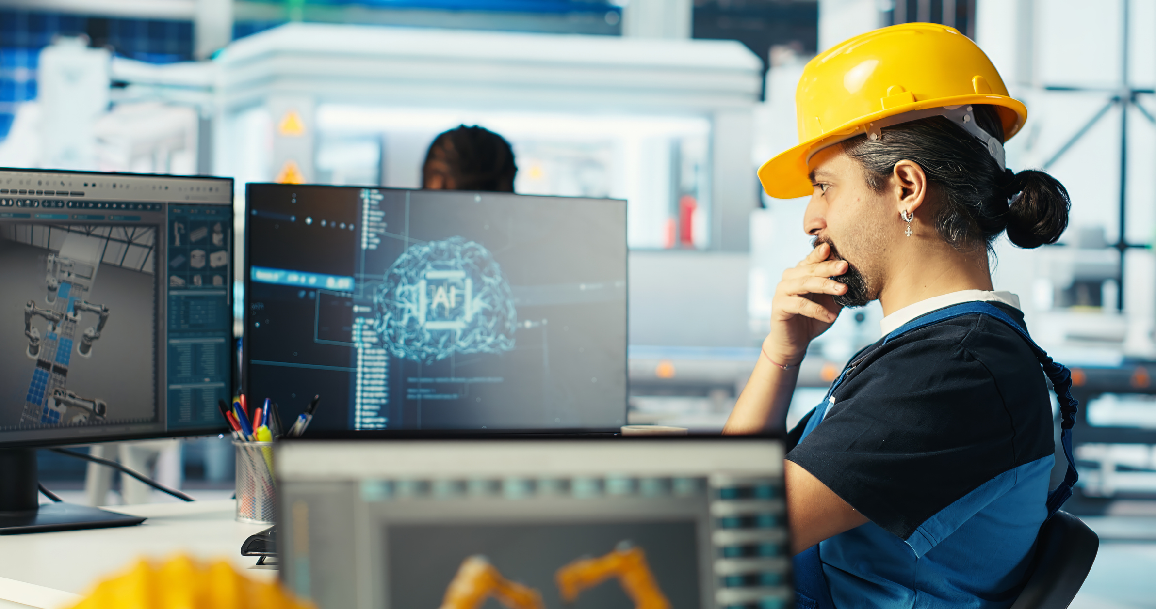 Engineer in yellow hard hat working at desk with dual monitors displaying AI technology interface in modern industrial facility