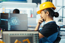 Engineer in yellow hard hat working at desk with dual monitors displaying AI technology interface in modern industrial facility