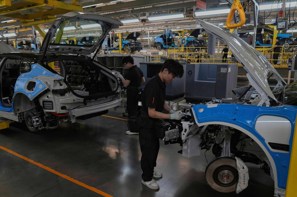 Workers assembling vehicles on a blue and white automotive manufacturing assembly line with industrial equipment overhead.