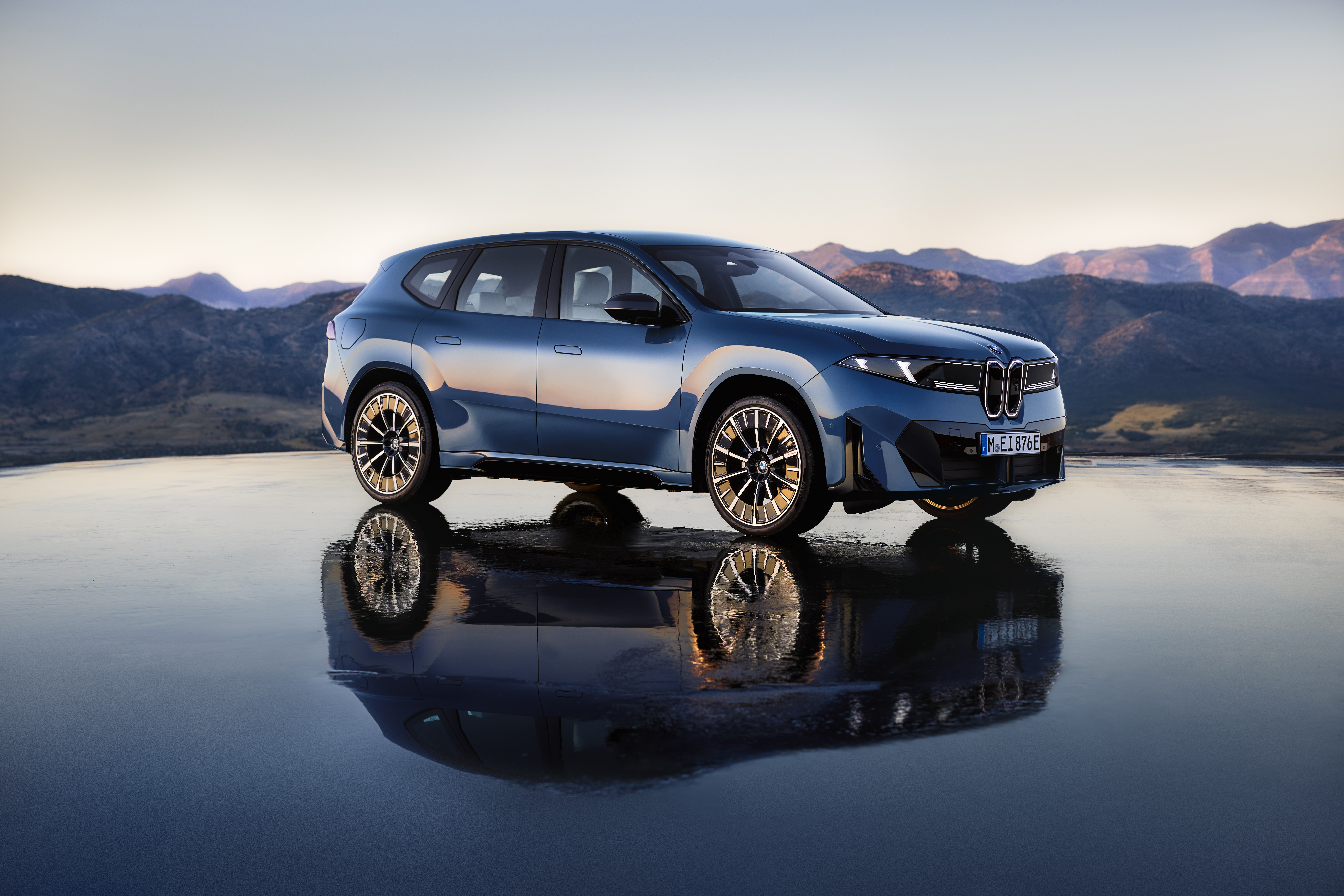 Blue and white BMW electric SUV displayed on reflective surface with mountain landscape backdrop