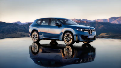Blue and white BMW electric SUV displayed on reflective surface with mountain landscape backdrop