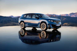 Blue and white BMW electric SUV displayed on reflective surface with mountain landscape backdrop