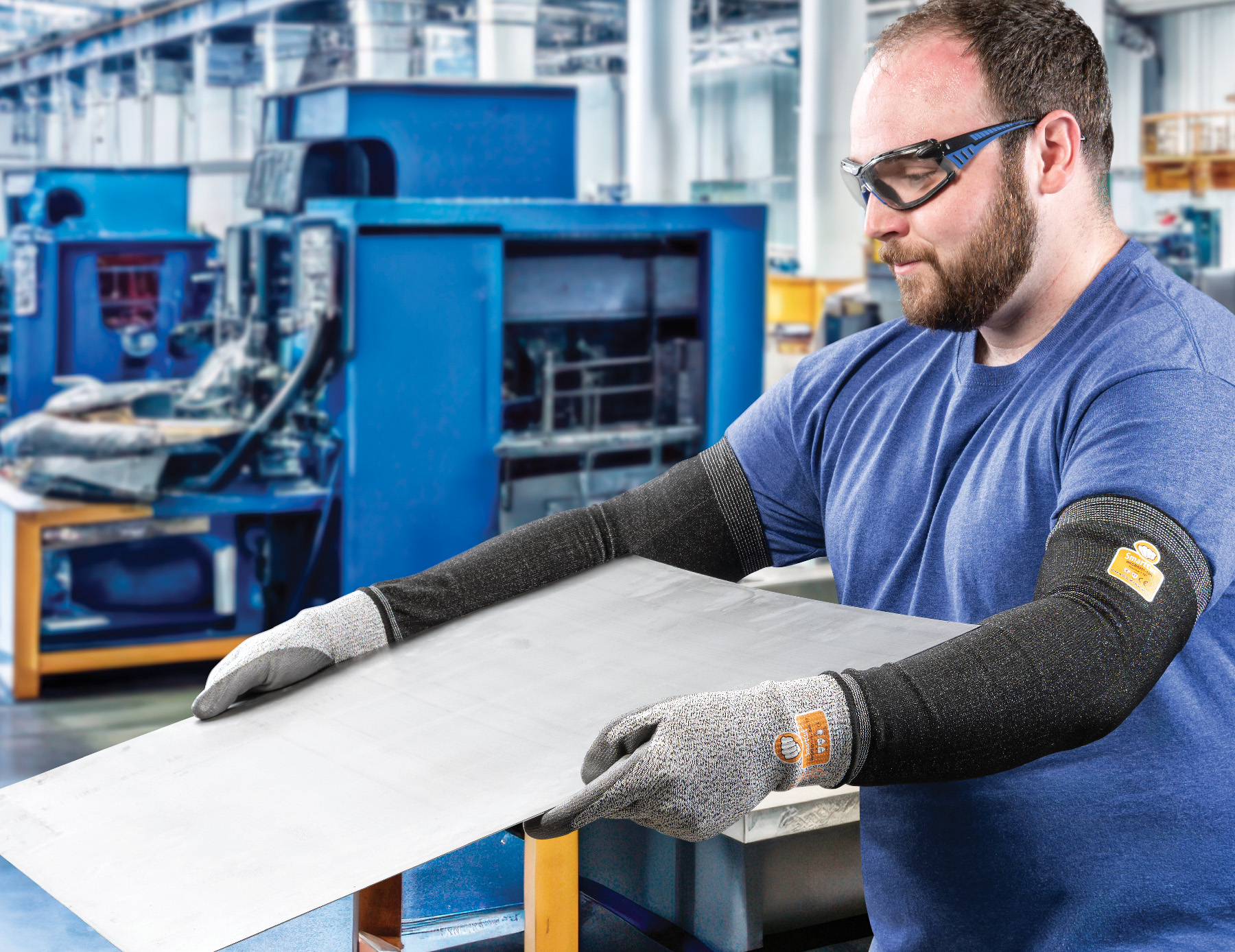 Factory worker in safety glasses and protective gloves inspecting white material at workstation with blue industrial machinery in background