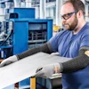 Factory worker in safety glasses and protective gloves inspecting white material at workstation with blue industrial machinery in background