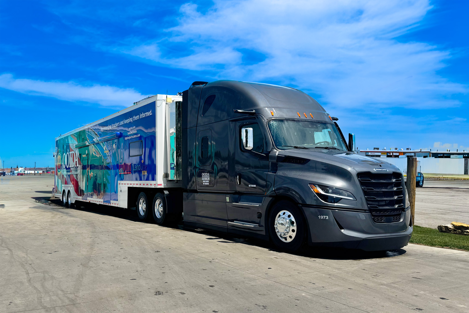 Black Freightliner semi-truck with colorful branded trailer parked on asphalt under blue sky