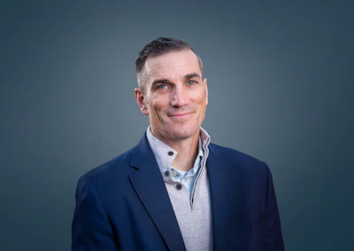 Professional headshot of a middle-aged man in navy blazer and light blue shirt against teal background