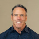 Professional headshot of a middle-aged man with gray hair wearing a dark blue shirt, smiling against a neutral background