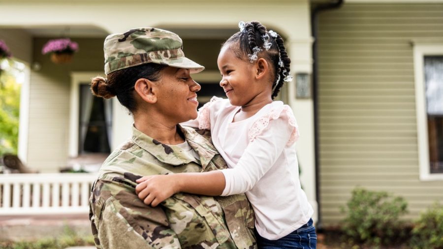 Military service member in camouflage uniform holding and smiling at a young child in front of a house