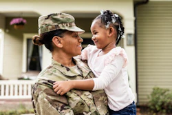 Military service member in camouflage uniform holding and smiling at a young child in front of a house