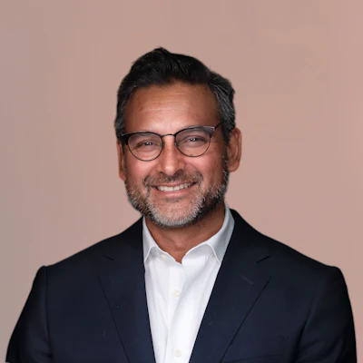 Professional headshot of a man wearing glasses and dark blazer with white shirt against pink background