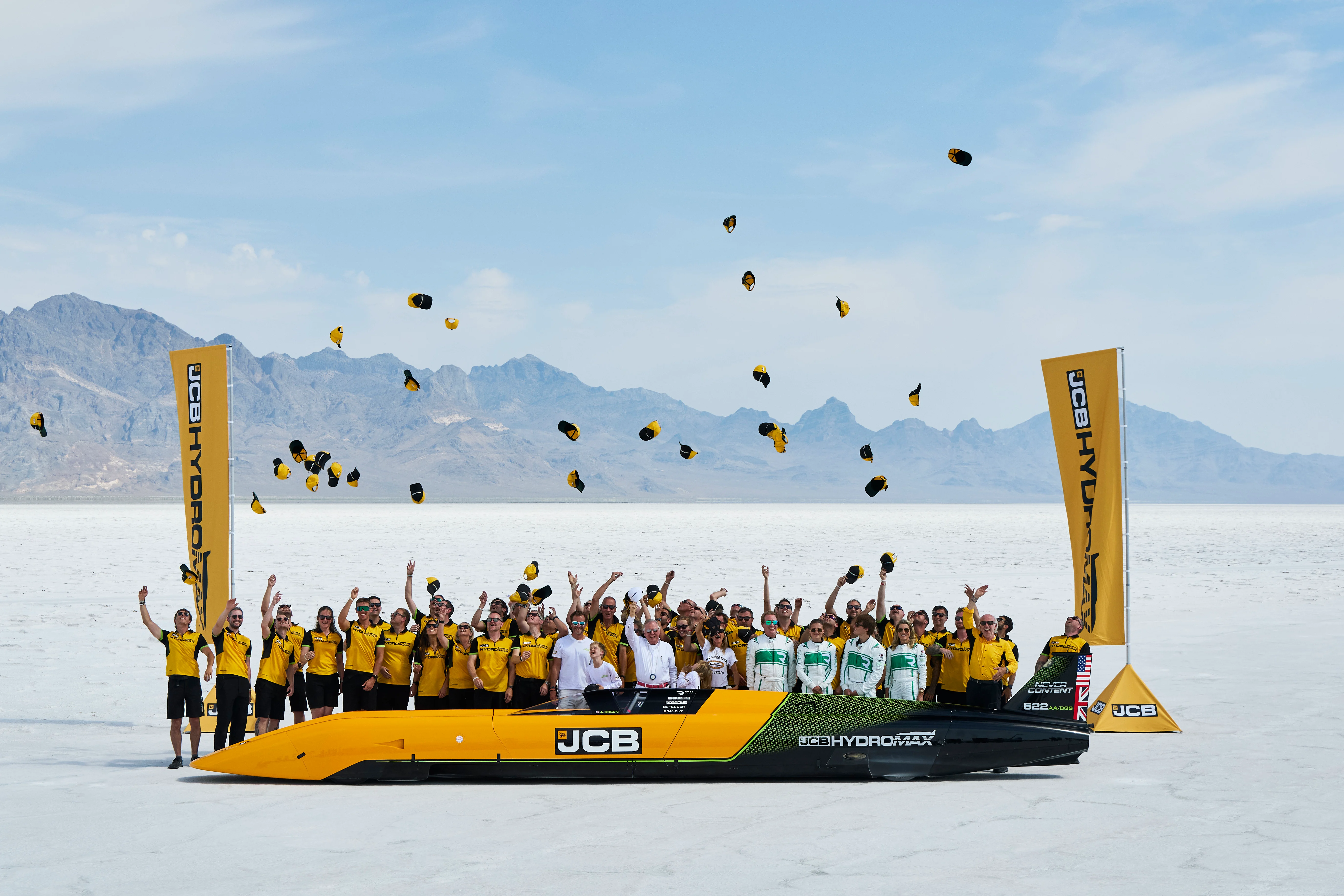Team celebrating on yellow JCB Hydromax vehicle in salt flat with mountains and hats thrown in air