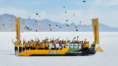 Team celebrating on yellow JCB Hydromax vehicle in salt flat with mountains and hats thrown in air