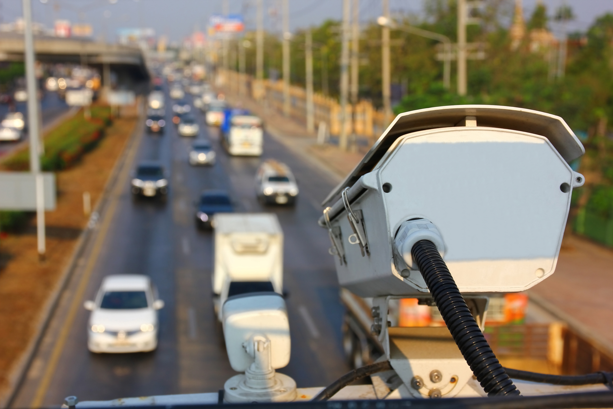 Traffic enforcement camera mounted on pole overlooking busy multi-lane highway with vehicles and urban infrastructure