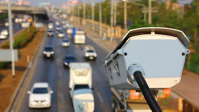 Traffic enforcement camera mounted on pole overlooking busy multi-lane highway with vehicles and urban infrastructure