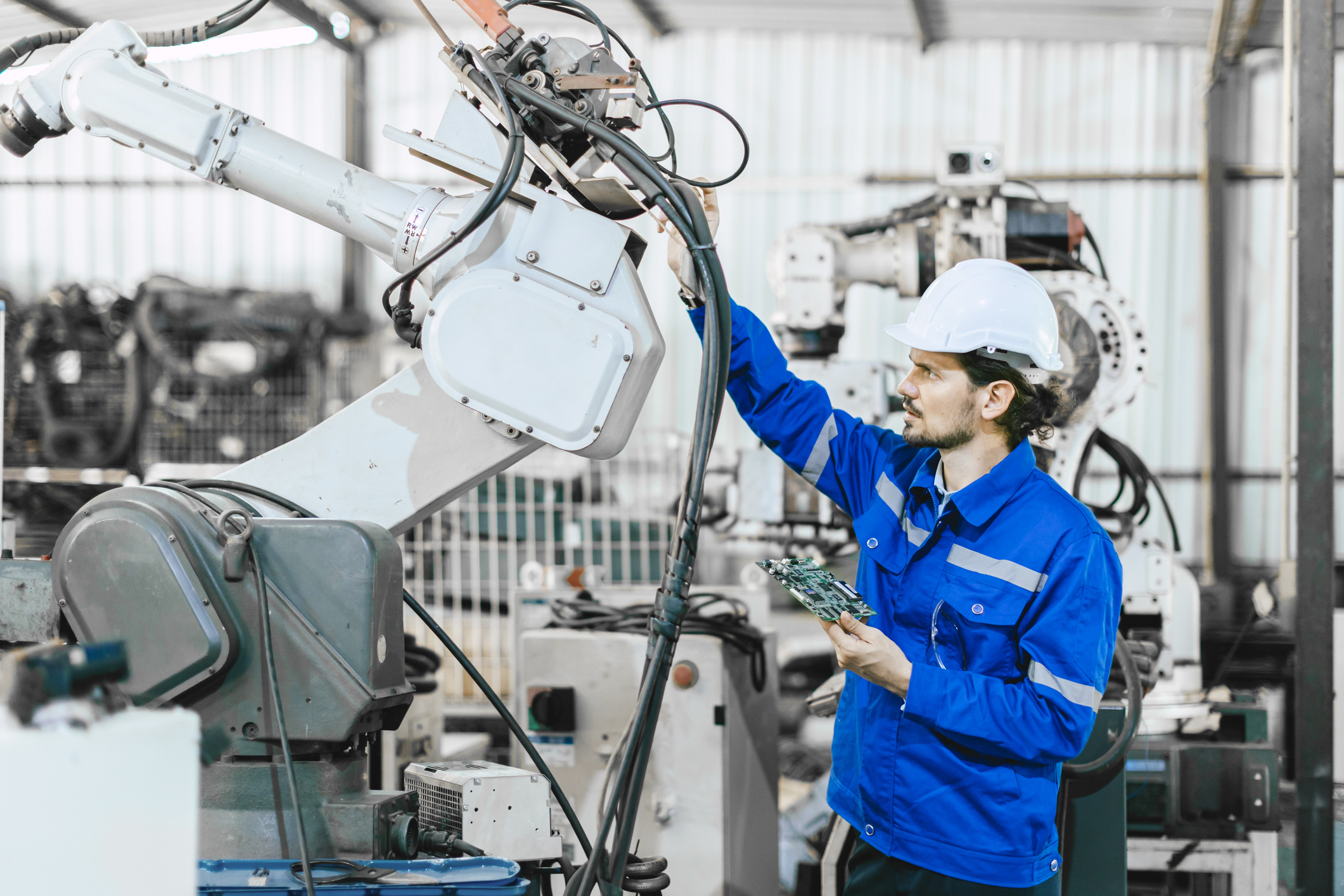 Technician in blue uniform and white hard hat inspecting industrial robotic arm equipment in manufacturing facility