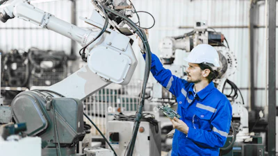 Technician in blue uniform and white hard hat inspecting industrial robotic arm equipment in manufacturing facility