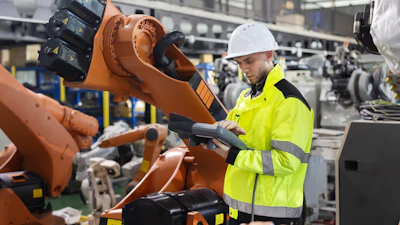 Factory worker in safety gear operating orange robotic arm in industrial manufacturing facility