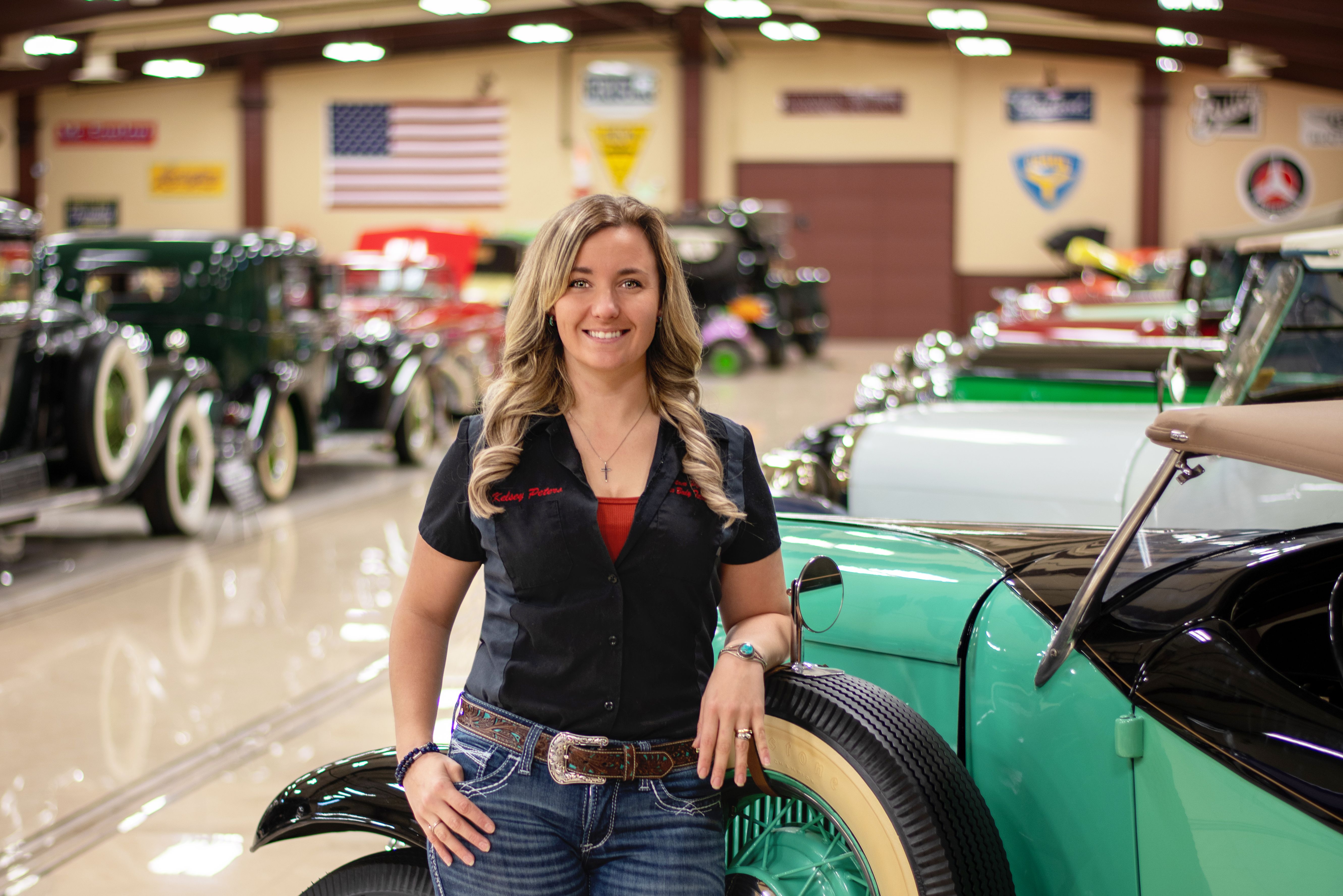 Woman in black vest smiling at vintage car show with classic vehicles displayed in garage