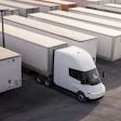White electric truck parked among white cargo trailers in a logistics yard