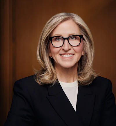 Professional headshot of woman wearing black blazer and glasses, smiling against brown background