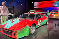 Vintage BMW race car with colorful red, green, and cyan livery displayed at automotive exhibition with spectator