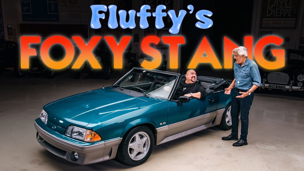Two men stand beside a teal and silver 1990 Ford Mustang convertible under a glowing 'Fluffy's Foxy Stang' neon sign in a garage setting.