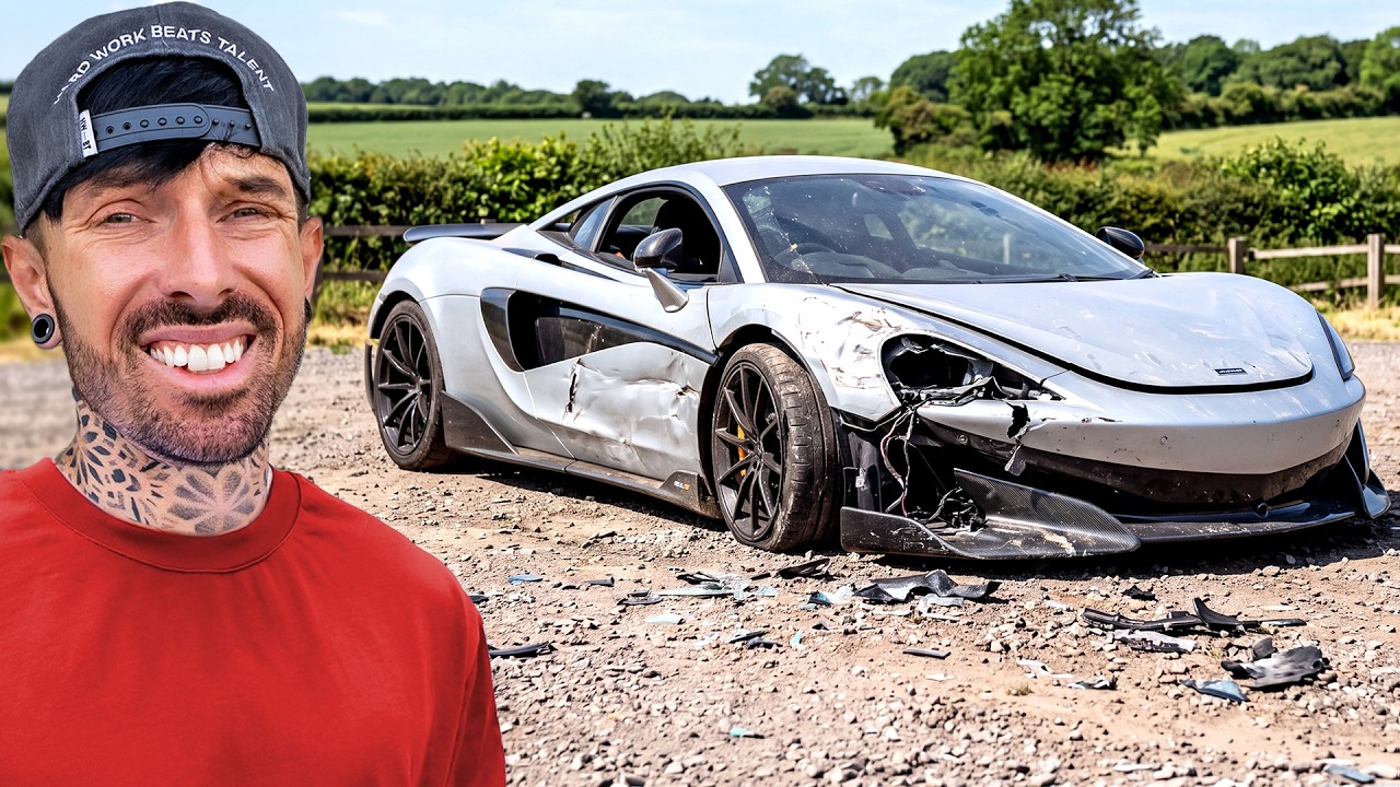 Man in red shirt smiling next to severely damaged white sports car in rural countryside setting