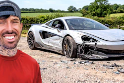 Man in red shirt smiling next to severely damaged white sports car in rural countryside setting