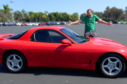 Man in green shirt posing on red sports car in parking lot with trees in background
