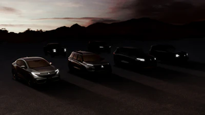 Multiple luxury vehicles parked at dusk with illuminated headlights against a mountain backdrop at sunset